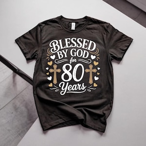 Puede incluir: Una camiseta negra de manga corta con texto gráfico en blanco y dorado que dice BLESSED BY GOD for 80 Years. El diseño incluye dos cruces doradas a ambos lados del número 80, rodeadas de pequeños iconos de corazones blancos y dorados.