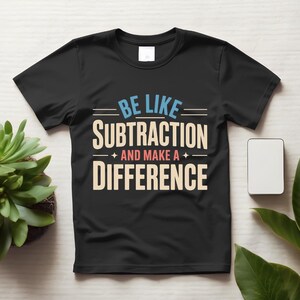 May include: A black short-sleeved t-shirt featuring the text "BE LIKE SUBTRACTION AND MAKE A DIFFERENCE" printed in light blue, cream, and salmon pink. The design includes horizontal lines and small star accents above and below the text.
