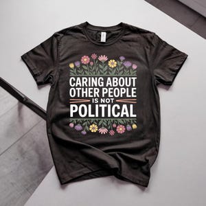 Könnte beinhalten: Ein schwarzes kurzärmeliges T-Shirt mit dem weißen Text "CARING ABOUT OTHER PEOPLE IS NOT POLITICAL" mittig auf der Brust. Der Text ist von farbenfrohen Illustrationen verschiedener Blumen und grüner Blätter ober- und unterhalb eingerahmt.