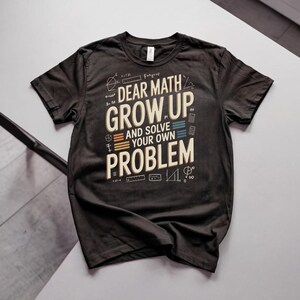 Pode incluir: Uma t-shirt preta de manga curta com o texto "DEAR MATH GROW UP AND SOLVE YOUR OWN PROBLEM" em creme e azul claro. O design inclui vários símbolos matemáticos brancos, formas geométricas e pequenas ilustrações de calculadoras.
