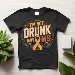 May include: A black short-sleeved t-shirt featuring a circular graphic with the text I'M NOT DRUNK I HAVE MS in orange and white lettering. The design includes an orange awareness ribbon centered below the text, surrounded by a sunburst pattern.