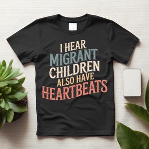 Könnte beinhalten: Ein schwarzes Kurzarm-T-Shirt mit dem Text I HEAR MIGRANT CHILDREN ALSO HAVE HEARTBEATS, der in einer mehrfarbigen, abgenutzten Schriftart gedruckt ist. Die Wörter sind in Weiß, Hellblau, Creme und Lachrosa auf der Brust angeordnet.