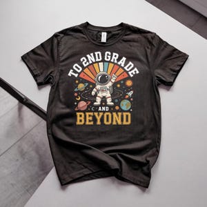 Puede incluir: Una camiseta negra de manga corta con un gráfico de un astronauta en el espacio con planetas, estrellas y un cohete. El texto TO 2ND GRADE AND BEYOND aparece en letras de bloque blancas y amarillas con un diseño de rayos de sol de estilo retro.