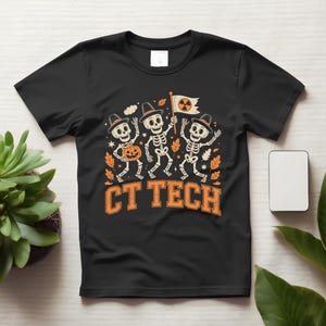 Könnte beinhalten: Ein schwarzes Kurzarm-T-Shirt mit einer Grafik von drei Skeletten mit Hüten, von denen eines einen Kürbis und ein anderes eine Flagge mit einem Strahlensymbol hält. Der Text CT TECH ist in Orange unter den Skeletten mit Herbstblättern gedruckt.