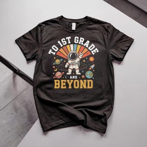Puede incluir: Una camiseta negra de manga corta con un gráfico de un astronauta en el espacio con planetas, estrellas y un cohete. El texto TO 1ST GRADE AND BEYOND aparece en letras blancas y amarillas con un diseño de rayos de sol de estilo retro.
