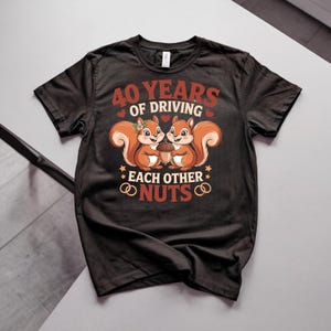Op de afbeelding: Een zwart t-shirt met korte mouwen en een afbeelding van twee eekhoorns die een eikel vasthouden. De tekst luidt 40 YEARS OF DRIVING EACH OTHER NUTS. Het ontwerp bevat kleine rode hartjes, gele sterren en twee gouden ringen in rood en geel.