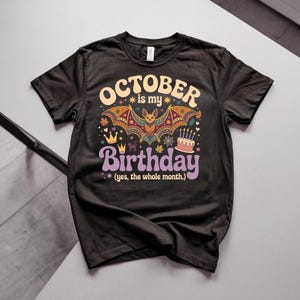 May include: A black short-sleeve t-shirt featuring a graphic with the text "OCTOBER is my Birthday (yes, the whole month.)". The design includes a colorful bat, two crowns, a small cat, a spider web, and a birthday cake with lit candles.
