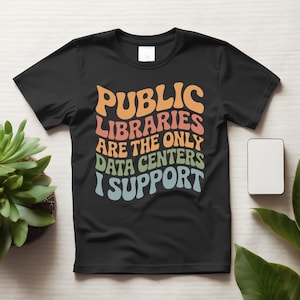 May include: A black short-sleeved t-shirt featuring the text "PUBLIC LIBRARIES ARE THE ONLY DATA CENTERS I SUPPORT" in a wavy, multi-colored font with orange, pink, green, and light blue letters centered on the chest area of the garment.