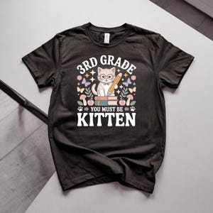 Puede incluir: Una camiseta negra de manga corta con el gráfico de un gatito con gafas y pajarita, sosteniendo un lápiz sobre libros. El texto dice 3RD GRADE YOU MUST BE KITTEN, rodeado de ilustraciones de mariposas, setas y hojas decorativas.