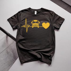 May include: A black short-sleeved t-shirt featuring a yellow graphic print. The design displays a heartbeat line that transitions into a yellow school bus icon, followed by a yellow heart shape, centered on the chest of the garment.