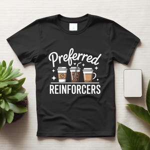 Peut inclure: Un t-shirt noir à manches courtes avec une impression graphique blanche. Le motif comprend le texte "Preferred REINFORCERS" en police grasse, centré au-dessus et au-dessous de trois tasses à café illustrées avec de petites étoiles.