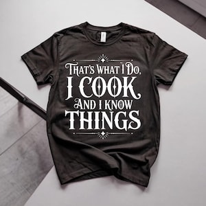 Puede incluir: Una camiseta negra de manga corta con texto blanco que dice: "THAT'S WHAT I DO, I COOK AND I KNOW THINGS". El diseño incluye detalles decorativos en forma de estrella encima y debajo del texto, centrado en la parte frontal de la prenda.