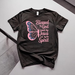 May include: A black short-sleeved t-shirt featuring a pink butterfly graphic on the left side. The text printed in pink reads: Blessed by God, Loved by Jesus, Led by the Spirit. The butterfly wings contain small blue circular accents.