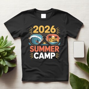 Könnte beinhalten: Ein schwarzes Kurzarm-T-Shirt mit einem Grafikdruck von Flieger-Sonnenbrillen, die im einen Glas eine Surfszene und im anderen einen tropischen Sonnenuntergang zeigen. Der Text 2026 SUMMER CAMP ist in Gelb, Orange und Weiß auf das T-Shirt gedruckt.