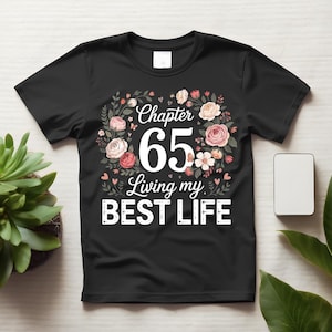 Pode incluir: Uma t-shirt preta de manga curta com um gráfico de coroa de flores que rodeia o texto "Chapter 65 Living my BEST LIFE" em branco e rosa. O design inclui várias rosas cor-de-rosa e brancas, folhas verdes e pequenos detalhes de corações.