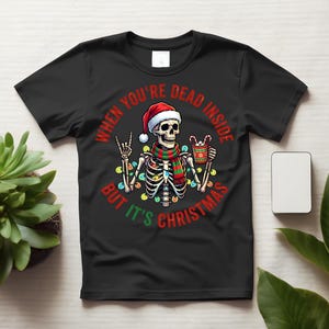 May include: A black short-sleeve t-shirt featuring a graphic of a skeleton wearing a Santa hat and a red and green striped scarf. The skeleton holds a mug with candy canes and is surrounded by colorful Christmas lights with the text WHEN YOU'RE DEAD INSIDE BUT IT'S CHRISTMAS.