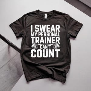 Könnte beinhalten: Schwarzes T-Shirt mit dem weißen Text "I Swear My Personal Trainer Can't Count". Eine Hantel-Grafik befindet sich zwischen "Trainer" und "Can't". Das T-Shirt hat einen Rundhalsausschnitt.