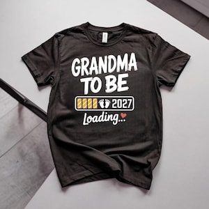 May include: A black short-sleeved t-shirt featuring the white text GRANDMA TO BE above a loading bar graphic with baby footprints and the year 2027, followed by the word Loading and a small red heart icon printed on the front of the garment.