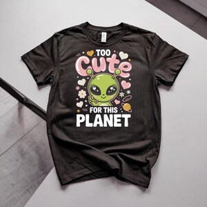 May include: A black short-sleeved t-shirt featuring a graphic of a green alien making a peace sign. The text reads "TOO CUTE FOR THIS PLANET" in white and pink letters, surrounded by small white hearts, flowers, and a small orange planet icon.