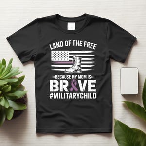 Op de afbeelding: Een zwart t-shirt met korte mouwen en een opdruk met de tekst LAND OF THE FREE, BECAUSE MY MOM IS BRAVE en #MILITARYCHILD. Het ontwerp bevat een gestileerde Amerikaanse vlag, een militaire laars en een paars bewustzijnslintje.