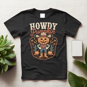 Könnte beinhalten: Ein schwarzes Kurzarm-T-Shirt mit einer Grafik eines kürbisköpfigen Charakters, der einen Cowboyhut, ein Halstuch und Stiefel trägt. Der Text "HOWDY Pumpkin" steht darüber, umrahmt von Illustrationen von Kakteen und Hufeisen.