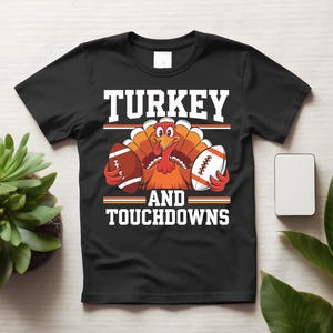 May include: A black short-sleeved t-shirt featuring a graphic of a cartoon turkey holding two American footballs. The text reads TURKEY AND TOUCHDOWNS in white block letters, accented by orange and white horizontal lines above and below the turkey.