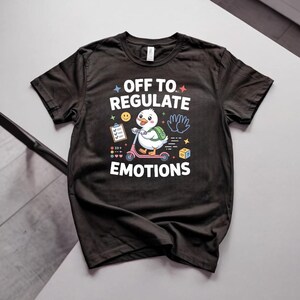 May include: A black short-sleeved t-shirt featuring a graphic of a white duck riding a pink scooter with a green backpack. The text reads OFF TO REGULATE EMOTIONS, accompanied by icons of a clipboard, a smiley face, hands, and a yellow block.