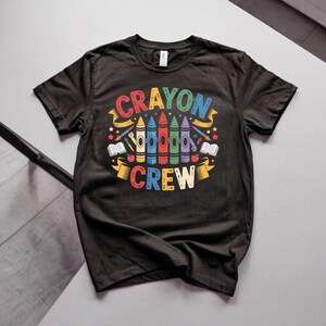 May include: A black short-sleeved t-shirt featuring a colourful graphic print with the text CRAYON CREW. The design includes five stylised crayons in yellow, blue, red, green, and purple, surrounded by small stars, open books, and paintbrushes.