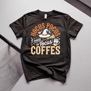 May include: A black short-sleeved t-shirt featuring a graphic print in orange, white, and black. The design includes a witch hat, a broom, a coffee cup with a pumpkin, and the text HOCUS POCUS I need to Focus COFFES with scattered coffee beans.
