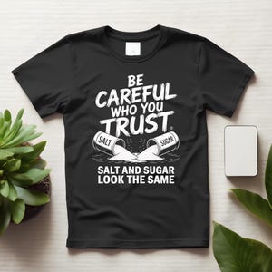 以下が含まれることがあります： BE CAREFUL WHO YOU TRUST. SALT AND SUGAR LOOK THE SAMEという白い文字がプリントされた黒い半袖Tシャツ。SALTとSUGARと書かれた2つの倒れた容器から白い粉が表面にこぼれ落ちている様子が描かれています。