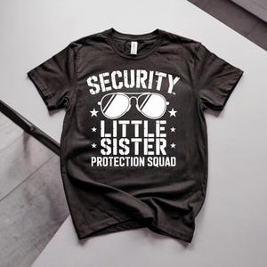 Puede incluir: Una camiseta negra de manga corta con texto gráfico en blanco que dice SECURITY, LITTLE SISTER y PROTECTION SQUAD. El diseño incluye una ilustración central de gafas de estilo aviador y cuatro pequeñas estrellas dispuestas alrededor del texto.