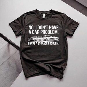 Puede incluir: Una camiseta negra de manga corta con texto blanco que dice: NO, I DON'T HAVE A CAR PROBLEM. I HAVE A STORAGE PROBLEM. Debajo del texto hay una ilustración gráfica de cuatro modelos de coches blancos diferentes alineados horizontalmente.
