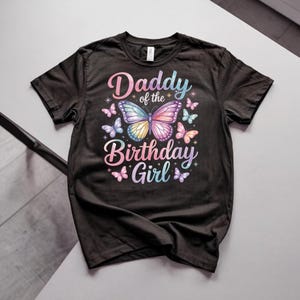 Könnte beinhalten: Ein schwarzes Kurzarm-T-Shirt mit einer großen Schmetterlingsgrafik in Pastellfarben in der Mitte. Der Schriftzug Daddy of the Birthday Girl ist in einer verlaufenden Schreibschrift gehalten und von kleinen Schmetterlingen umgeben.