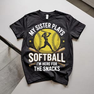 May include: A black short-sleeved t-shirt featuring a graphic of a softball player swinging a bat in front of a yellow softball. The text reads: MY SISTER PLAYS SOFTBALL I'M HERE FOR THE SNACKS. Two wooden baseball bats are crossed below the ball.