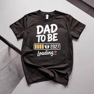 May include: A black short-sleeved t-shirt featuring white text that reads DAD TO BE. Below the text is a loading bar graphic with baby footprints and the year 2027, followed by the word Loading and a small red heart icon printed on the front.