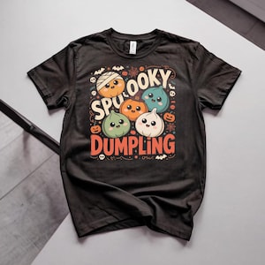 May include: A black short-sleeve t-shirt featuring a graphic print of four cute, smiling dumplings with Halloween-themed elements like a mummy wrap, a witch hat, and small pumpkins. The text reads SPOOKY DUMPLING in orange and cream colors.
