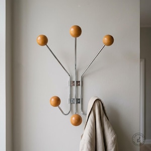 May include: A wall-mounted coat rack featuring a vertical chrome metal frame with five extending arms. Each arm terminates in a smooth, round, light brown wooden ball. A beige fabric garment hangs from one of the lower hooks on the right side.