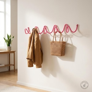 May include: A bright pink wall-mounted coat rack featuring a continuous, wavy, serpentine design with multiple hooks. A tan jacket hangs on the left side, and a woven tan tote bag with a grid pattern hangs from the right side of the rack.