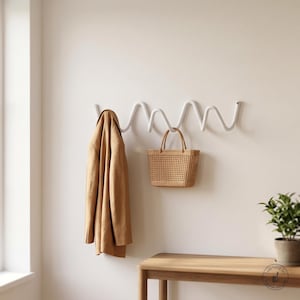 May include: A white wall-mounted coat rack with a continuous wavy, serpentine design. A tan jacket hangs from the left side of the rack, and a woven tan straw handbag with two top handles hangs from the centre of the wavy metal structure.