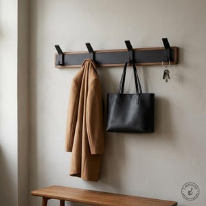 May include: A wall-mounted coat rack featuring a horizontal wooden base with a matte black centre panel and four matching black metal hooks. A tan coat hangs on the first hook, a black leather tote bag on the third, and keys on the fourth.