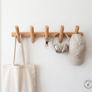 May include: A light-coloured wooden wall-mounted coat rack with five cylindrical pegs. Hanging from the pegs are a plain beige canvas tote bag, a metal keyring with keys, a pair of beige over-ear headphones, and a beige baseball cap.