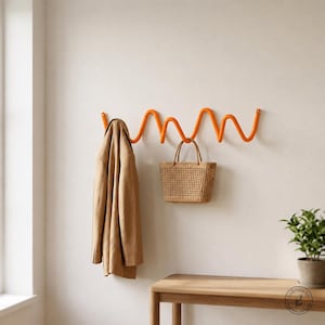 Könnte beinhalten: Eine leuchtend orangefarbene Wandgarderobe mit einem durchgehenden, wellenförmigen Design. Die Garderobe verfügt über mehrere Haken, die durch die Kurven des Metallrahmens gebildet werden. Ein beigefarbener Mantel hängt links, eine Strohtasche rechts.
