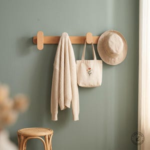 May include: A light wood wall-mounted coat rack with rounded ends. Hanging from the rack is a cream-coloured knit jumper, a cream canvas tote bag featuring a small embroidered floral design, and a beige wide-brimmed hat made of fabric.