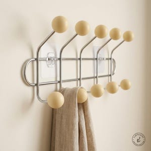 May include: A wall-mounted coat rack featuring a chrome metal frame with ten hooks, each topped with a smooth, round, cream-coloured wooden ball. The rack is secured to the wall with two clear adhesive plates and holds a beige linen cloth.