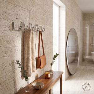 May include: A silver-toned metal wall-mounted coat rack featuring a continuous, undulating wave design. The rack is mounted on a beige stone wall and holds a beige knit scarf and a tan leather shoulder bag with a thin strap and gold hardware.