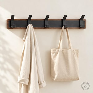 Può includere: Un appendiabiti da parete con una base orizzontale in legno scuro, un pannello in metallo nero opaco e cinque ganci in metallo nero abbinati. Una camicia beige chiaro e una borsa in tela beige abbinata sono appese ai ganci dell'appendiabiti.