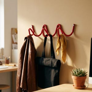 Könnte beinhalten: Eine an der Wand montierte rote Metall-Garderobenleiste mit durchgehendem Wellendesign und drei Haken. Eine braune Lederjacke hängt am linken Haken, eine schwarze Stofftasche am mittleren Haken und ein gelber Stoffschal am rechten Haken.