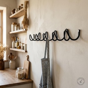Peut inclure: Un porte-manteau mural en métal noir présentant un design en spirale continu et répétitif avec plusieurs crochets. Un tablier à motifs bleus et blancs est suspendu à l'un des crochets. Le porte-manteau est fixé sur un mur texturé clair.
