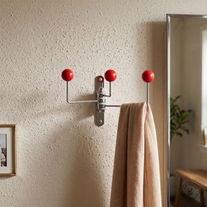 Puede incluir: Un perchero de pared con una base vertical de metal plateado y tres brazos ajustables, cada uno terminado en un pomo esférico de color rojo brillante. Una bufanda de tela beige cuelga sobre uno de los brazos del perchero de metal.