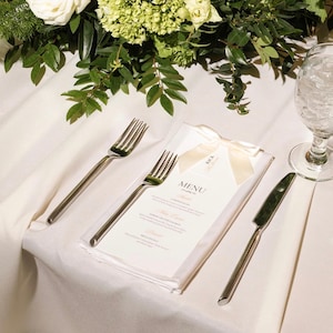 May include: A white paper menu card with the word MENU printed in black, adorned with a cream-colored satin ribbon bow. The menu is placed on a white tablecloth alongside two silver-toned forks, a silver-toned knife, and a glass of water.